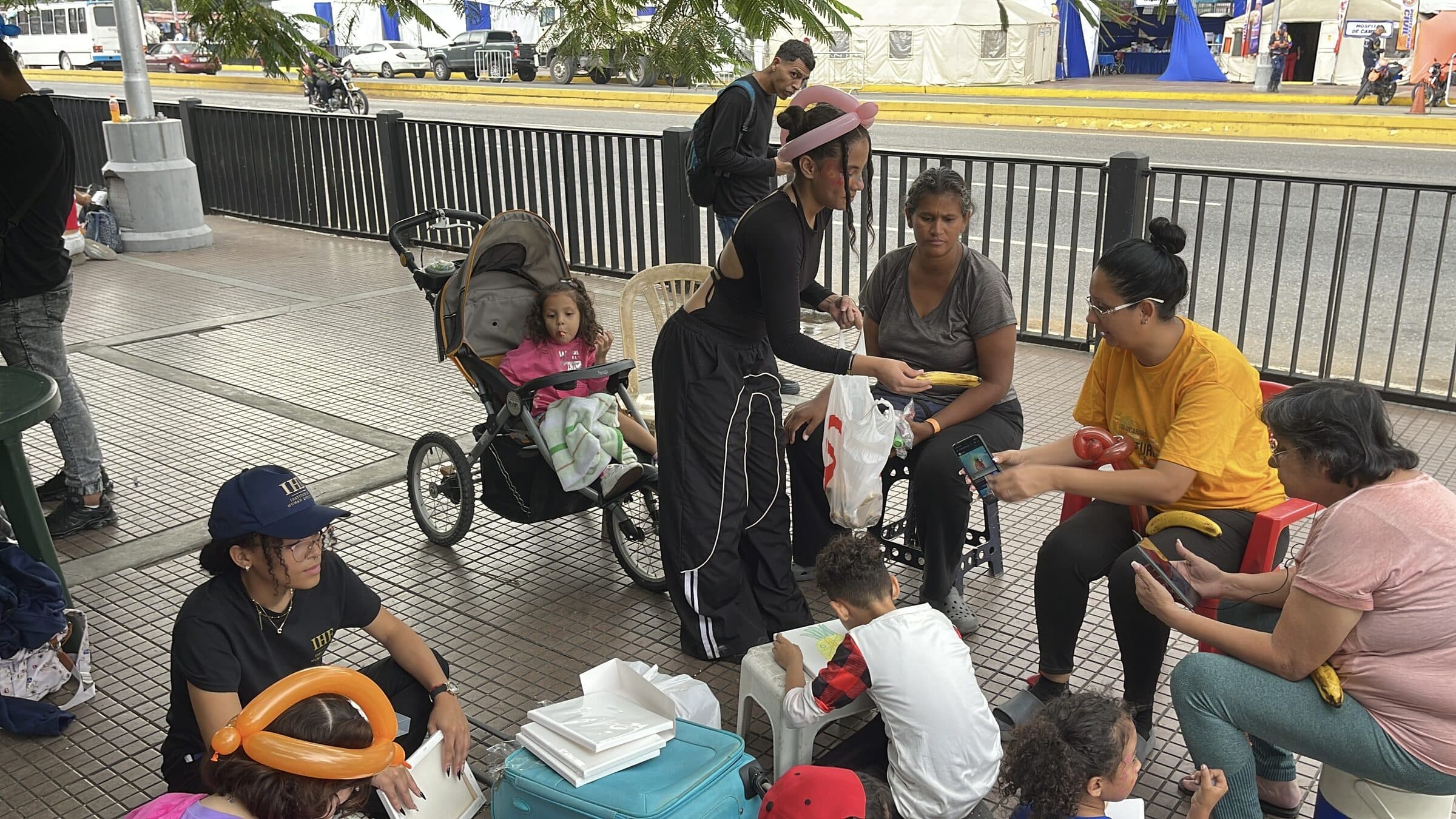 Una voluntaria entrega fruta fresca a una madre junto al cochecito de su hija; al fondo se ven las carpas del campamento al otro lado de la calle en Caracas.