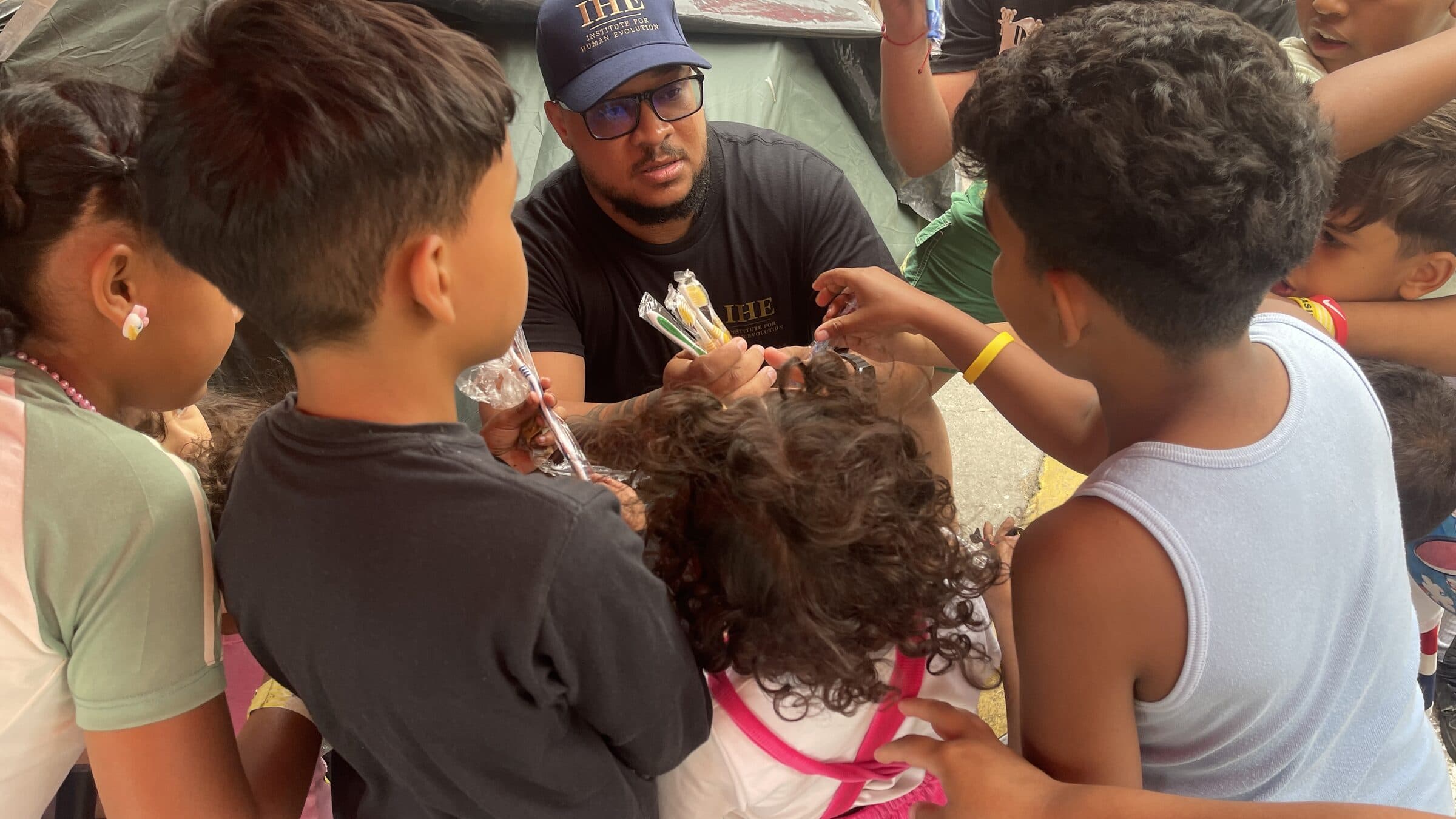 Una voluntaria de IHE entrega cepillos de dientes a niños que extienden las manos para recibirlos en el campamento.