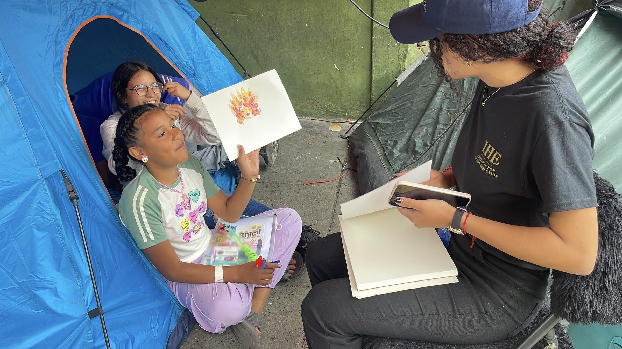 Una niña sonríe ampliamente mientras muestra su dibujo a la cámara durante un taller de arte de IHE.