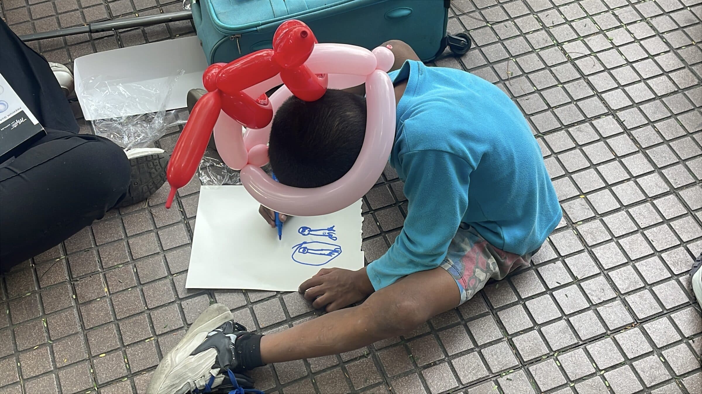 Un niño con un sombrero de globos se inclina sobre su dibujo en el suelo, completamente concentrado en colorear; su rostro no es visible.