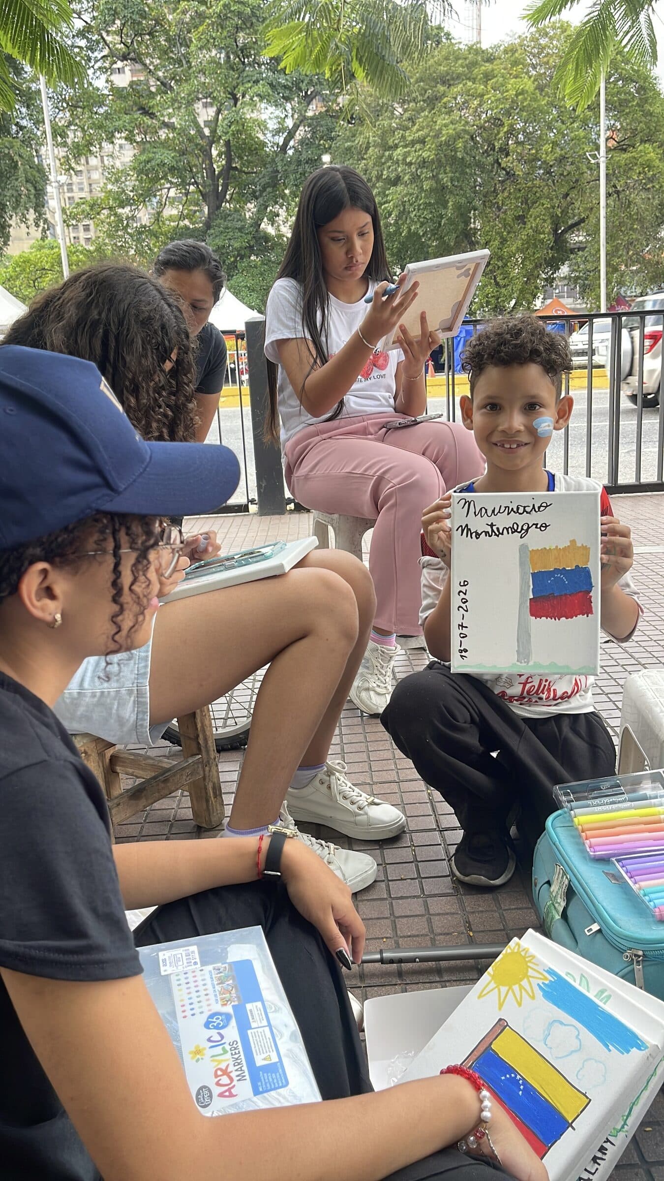 Un niño sonriente muestra a la cámara su cuadro firmado de la bandera de Venezuela en el taller de arte del campamento.