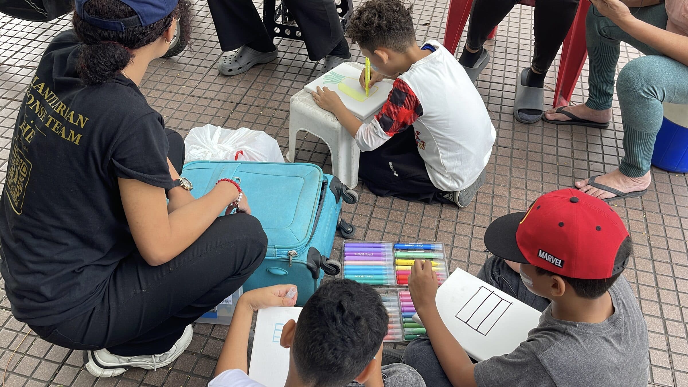 Un niño, de espaldas, pinta una piña en un pequeño lienzo durante un taller de arte de IHE; en primer plano, la camiseta del equipo humanitario de una voluntaria.