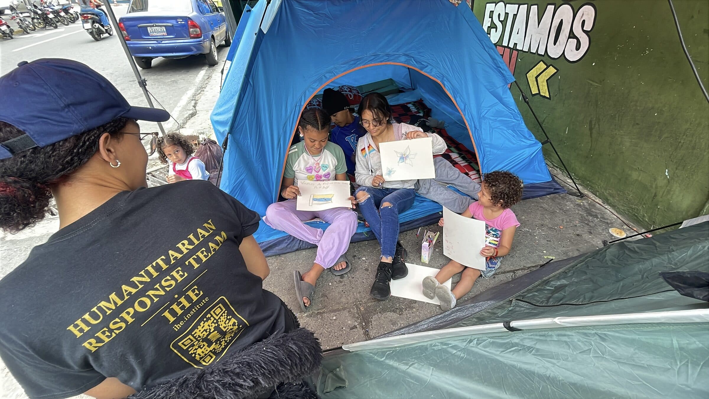 Niños muestran orgullosos sus dibujos terminados, entre ellos una bandera de Venezuela, junto a una voluntaria de IHE en el taller de arte del campamento.