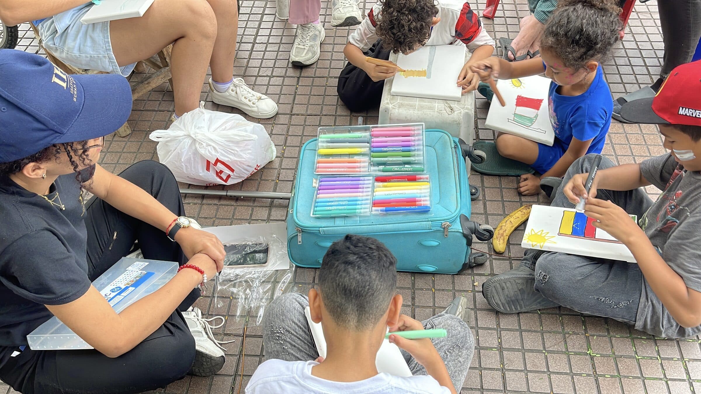 Niños pintan banderas, entre ellas la de Venezuela, en pequeños lienzos alrededor de una maleta usada como mesa llena de estuches de marcadores.