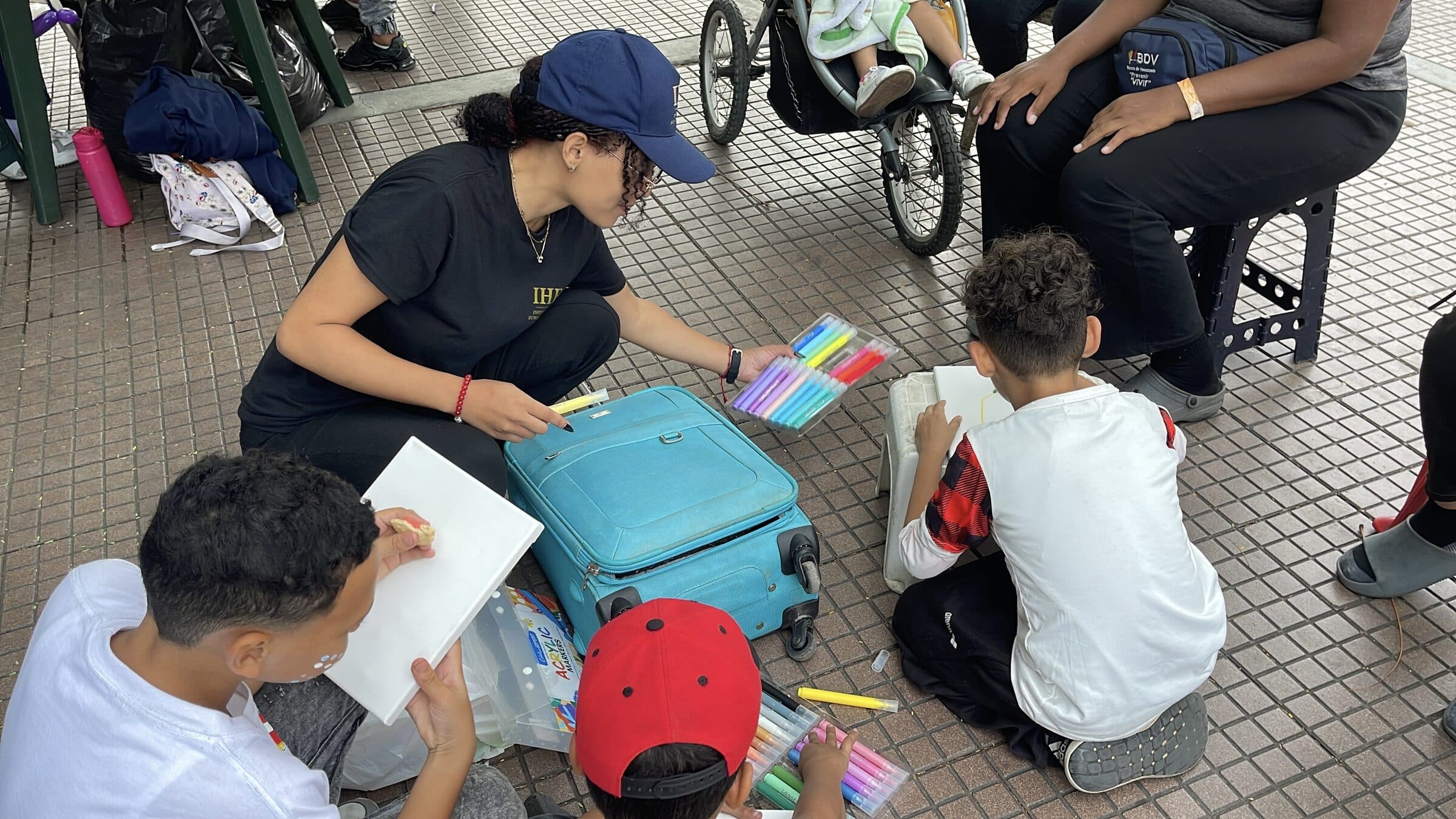 Una voluntaria de IHE ofrece una bandeja de marcadores a niños que pintan lienzos en un taller de arte al aire libre en un campamento de familias desplazadas en Venezuela.