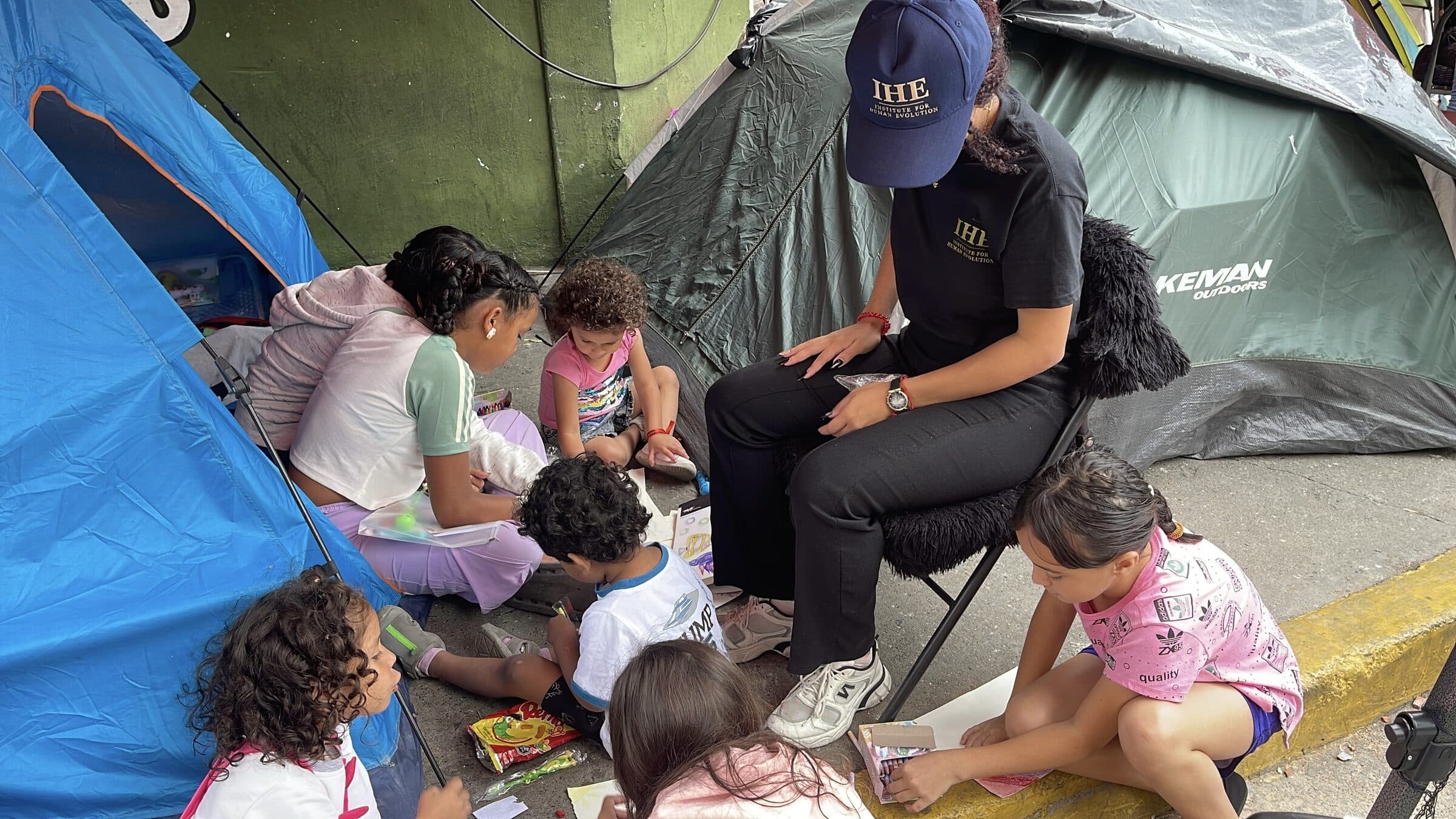 Una voluntaria de IHE sentada entre niños concentrados dibujando entre las carpas del campamento.
