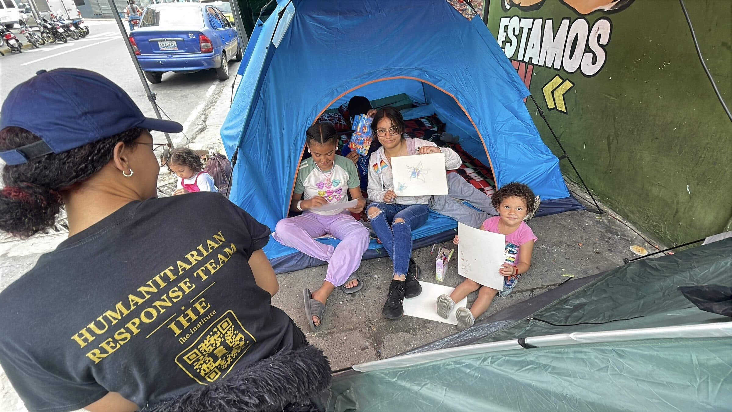 Vistas desde arriba, manos de niños dibujan sobre papel en el bordillo mientras una voluntaria de IHE los acompaña.
