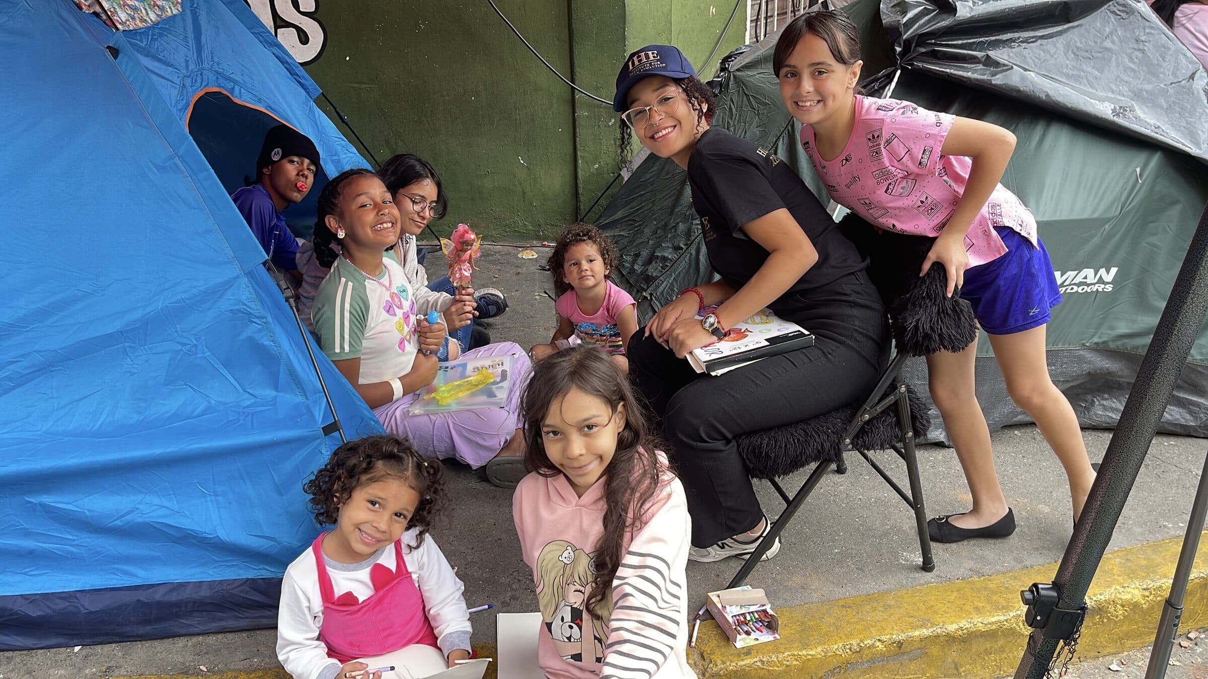 An IHE volunteer surrounded by smiling children in front of the tents, a spontaneous group portrait at the camp.