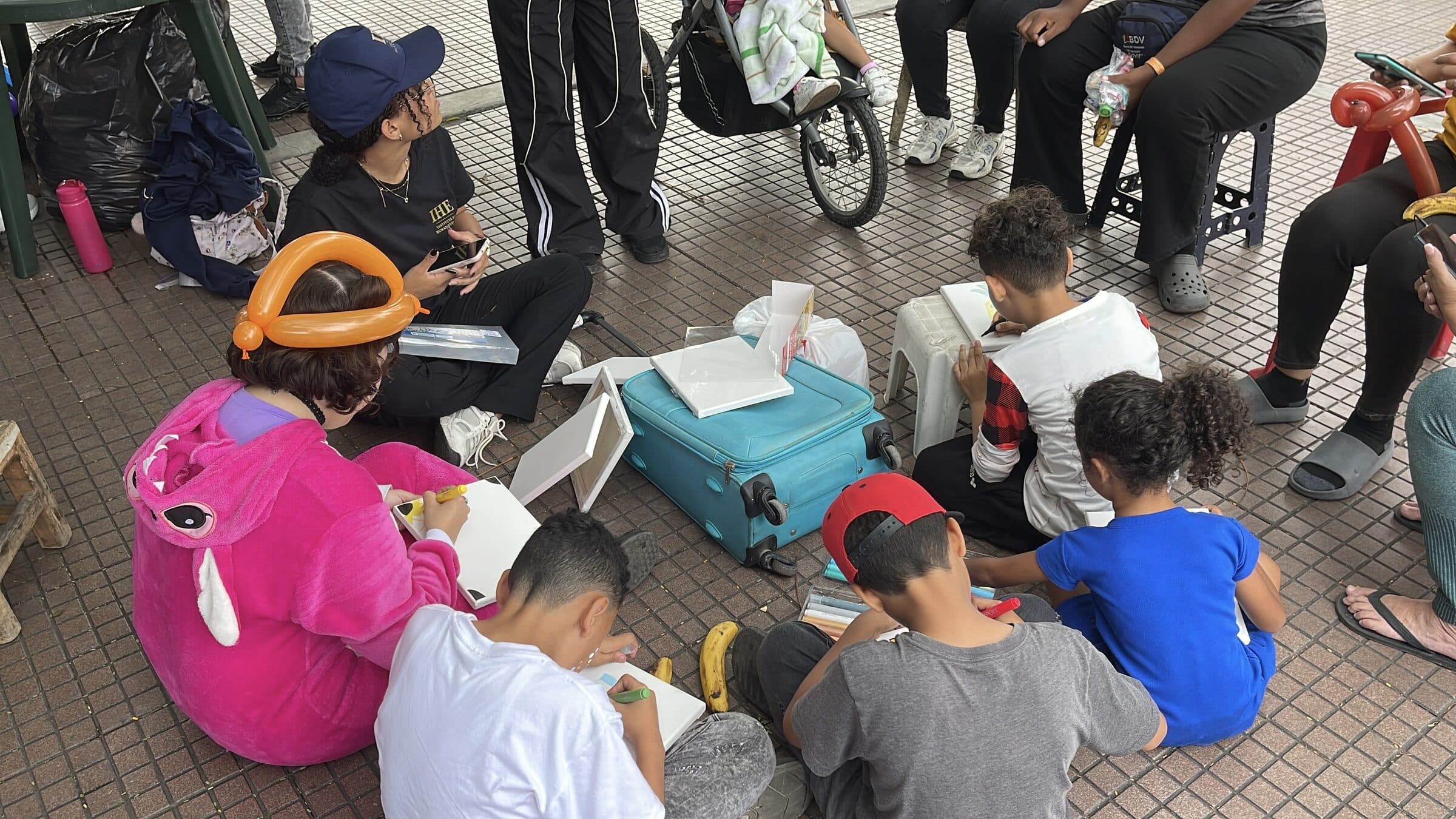 Plano amplio de un taller de arte al aire libre: niños de espaldas pintan lienzos alrededor de una maleta con materiales mientras las familias se reúnen en círculo.