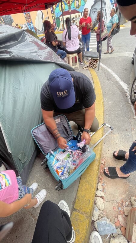 Juan kneeling beside a rolling suitcase, unpacking relief supplies near a tent.