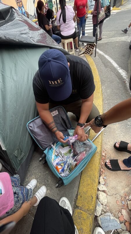 Juan kneeling beside a rolling suitcase, unpacking relief supplies near a tent.