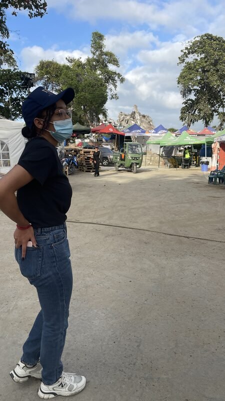 Cori, in a face mask, stands looking down a street lined with tents and stalls.